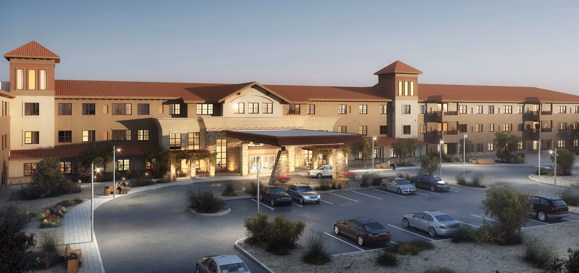 Exterior view of a large, three-story senior living facility building with a covered entrance and multiple parked cars in the parking lot during dusk. The building has a beige facade with red-tiled roofs and several windows, surrounded by landscaped bushes and street lamps.