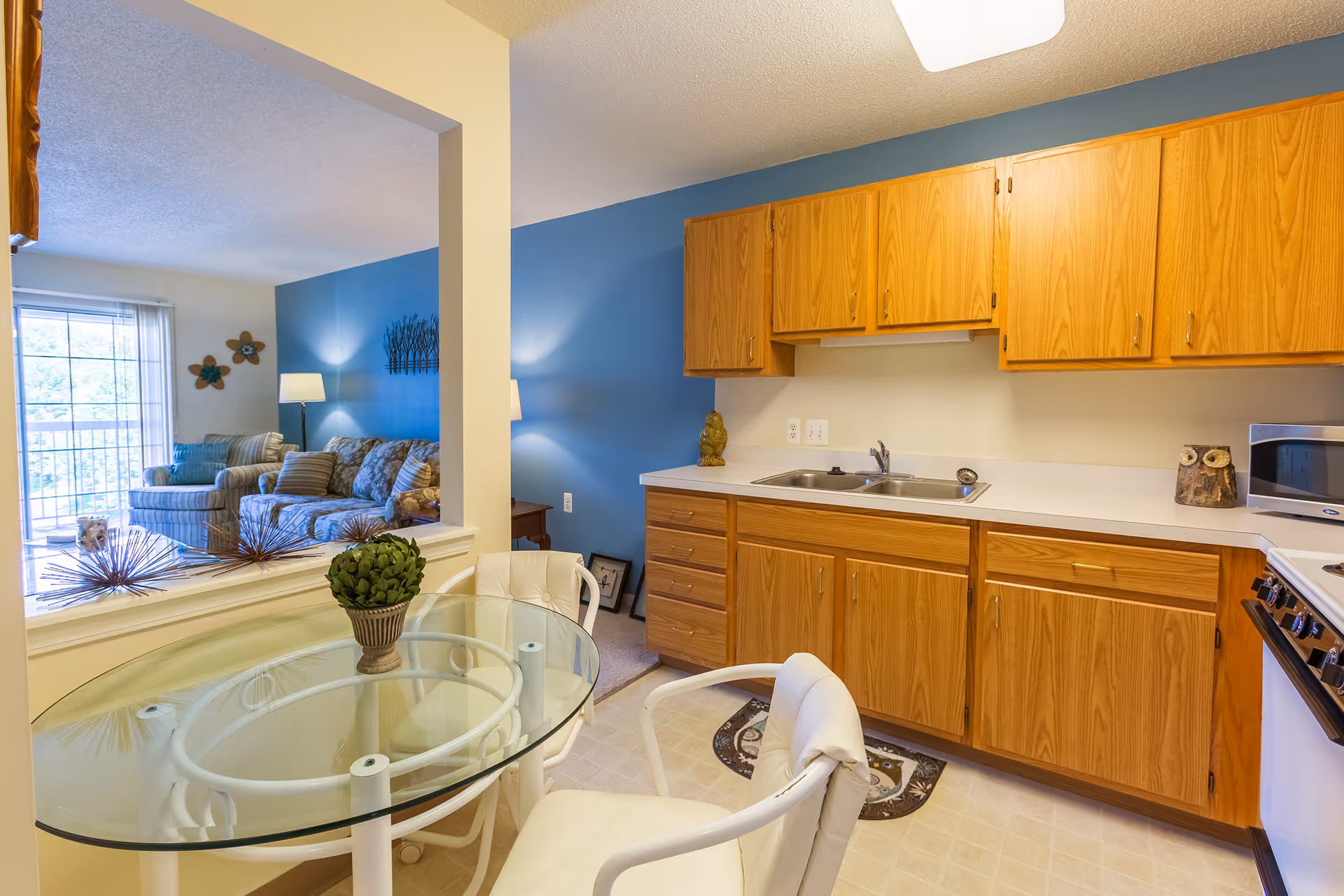 Interior view of a senior living facility apartment showing a small kitchen area with wooden cabinets, a double sink, a microwave, and a stove. In the foreground, there is a glass-top dining table with two white cushioned chairs. Beyond the kitchen, a living room with a blue accent wall, a sofa, an armchair, a floor lamp, and wall decorations is visible. A sliding glass door leads to an outdoor balcony.