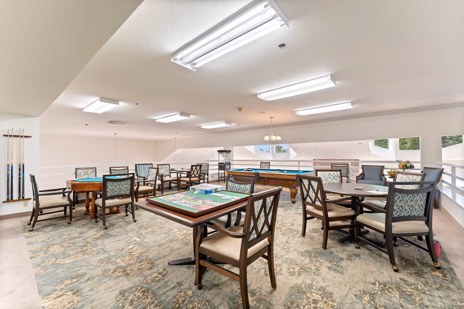 A spacious recreational room with multiple tables and chairs arranged for games and social activities. There is a puzzle on one table, a pool table with billiard balls, and a popcorn machine in the background. The room has large windows letting in natural light and a patterned carpet covering the floor.