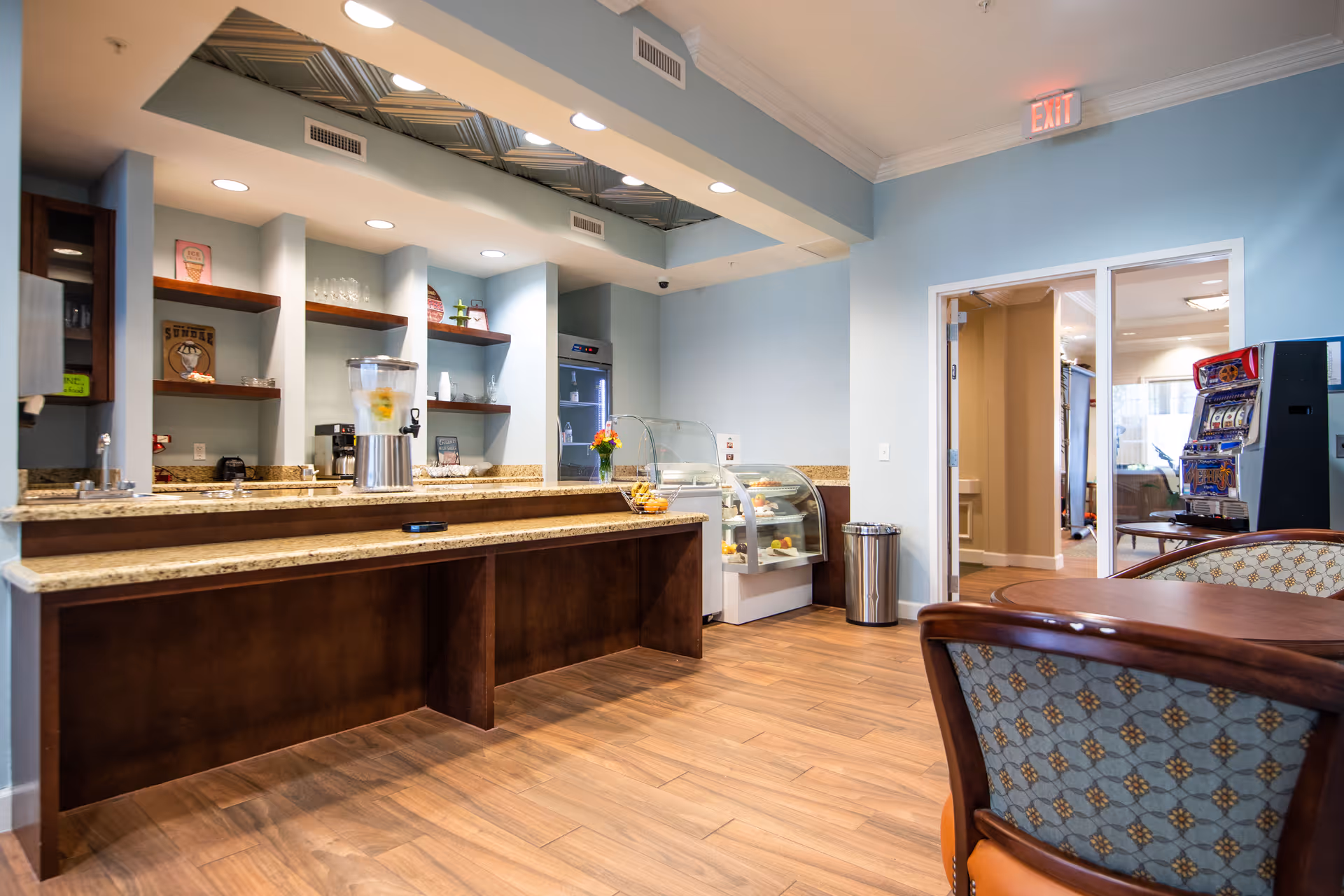 Interior view of a senior living facility's refreshment area with a long granite countertop, water dispenser, glass display cases with pastries, wooden shelves with decorative items, and a seating area with upholstered chairs. The room has light blue walls, wood flooring, and an exit sign above a doorway leading to another room.