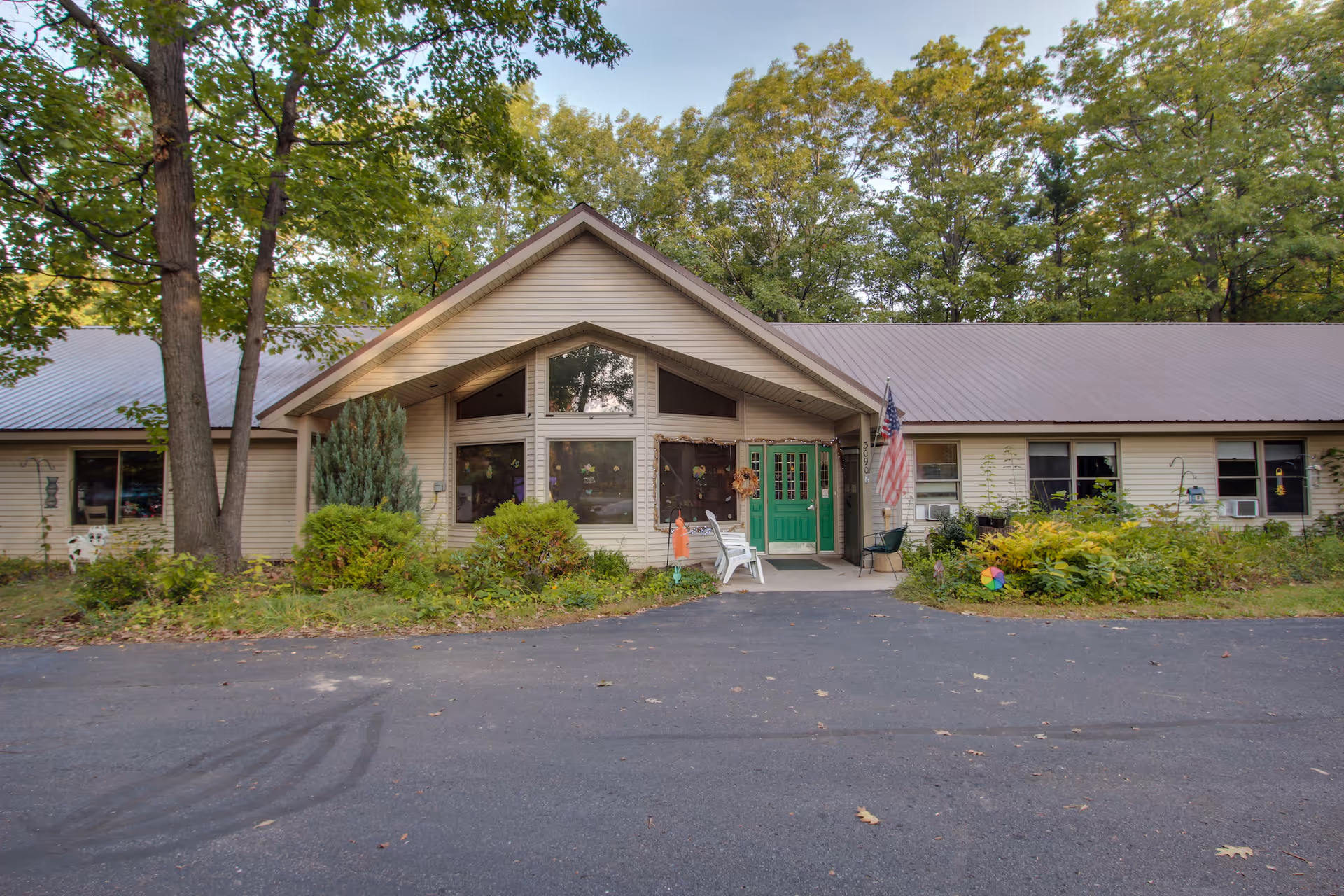 Front exterior view of a single-story building with beige siding and a green double door entrance. The building is surrounded by trees and bushes, with an American flag displayed near the entrance. There are two white chairs placed outside the door on a small concrete porch.