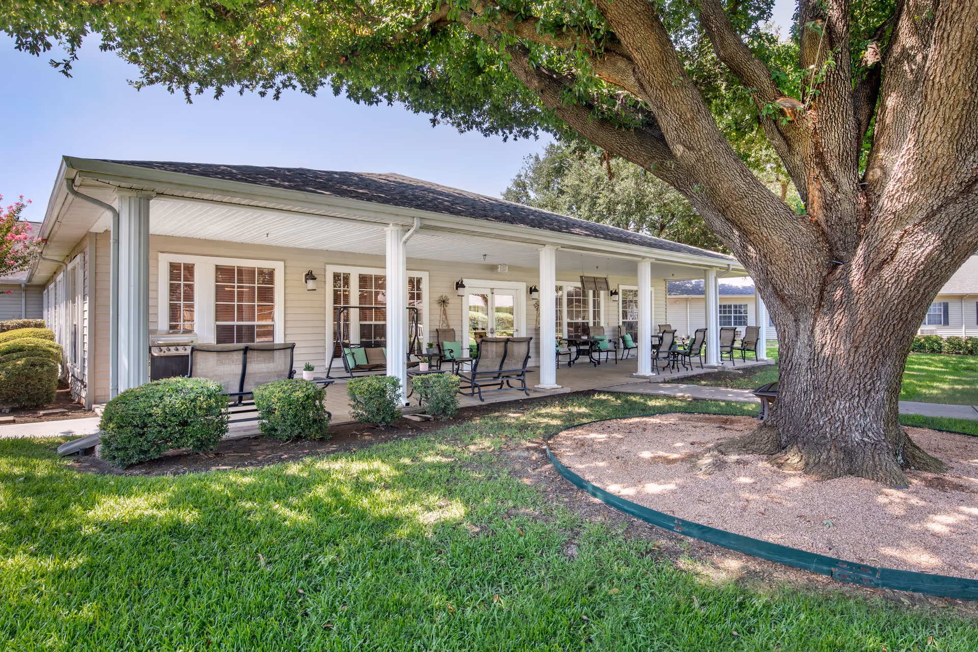 Outdoor covered patio area at Brookdale Tanglewood Oaks with multiple seating arrangements including chairs and benches. The patio is supported by white columns and overlooks a well-maintained lawn with a large tree in the foreground providing shade.