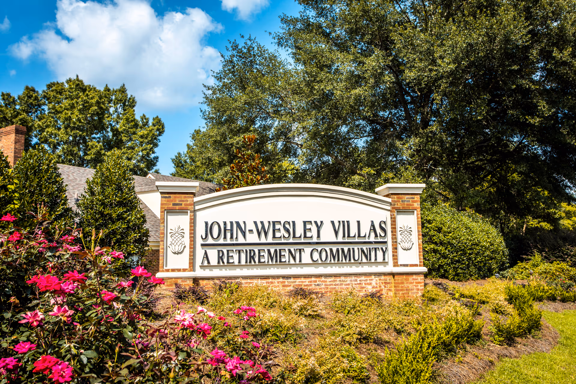 Entrance sign for John-Wesley Villas, a retirement community, surrounded by green bushes and pink flowers with trees and a partly cloudy sky in the background.