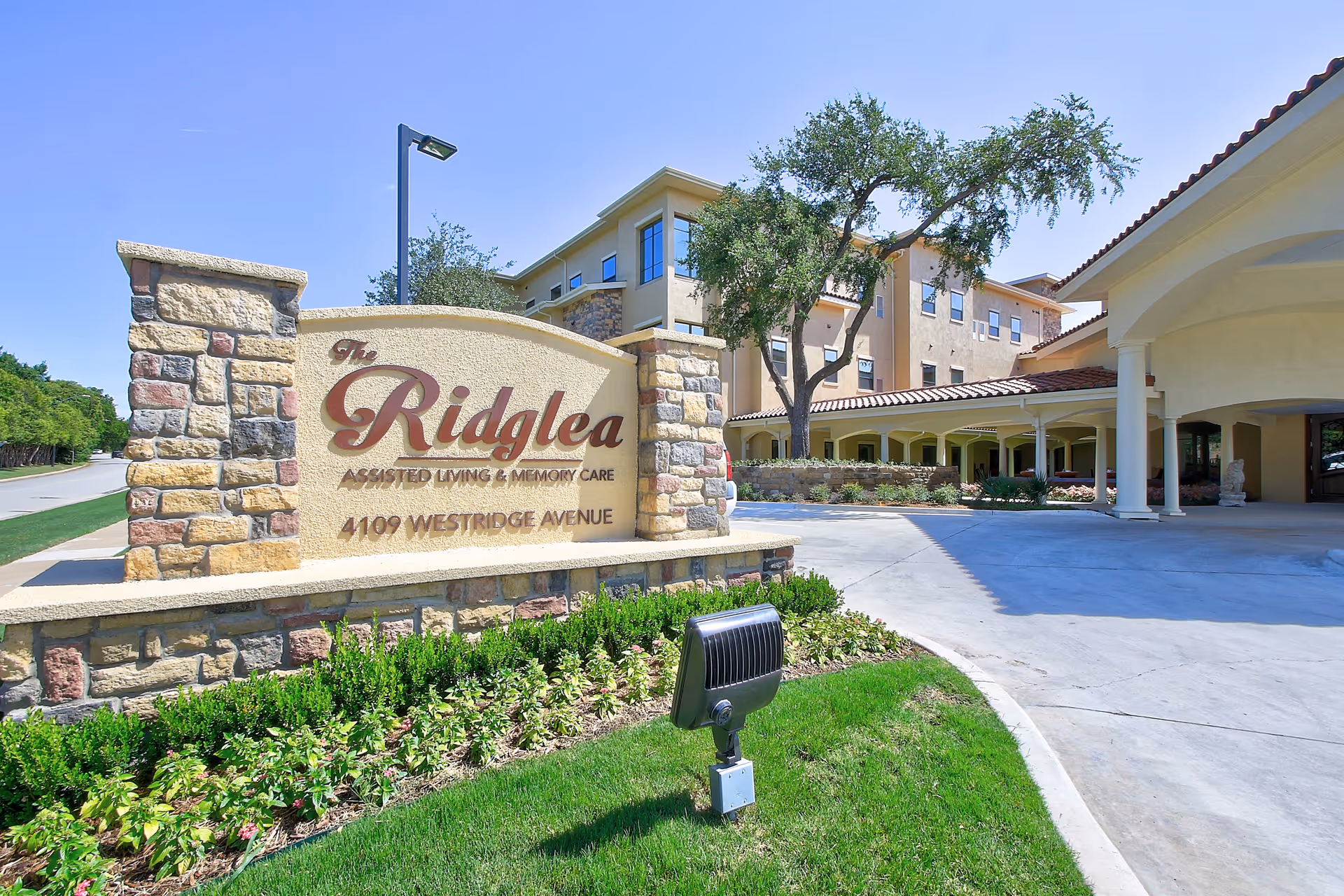 Stone sign reading "The Ridglea" in front of a senior living building with landscaping and a covered driveway entrance.