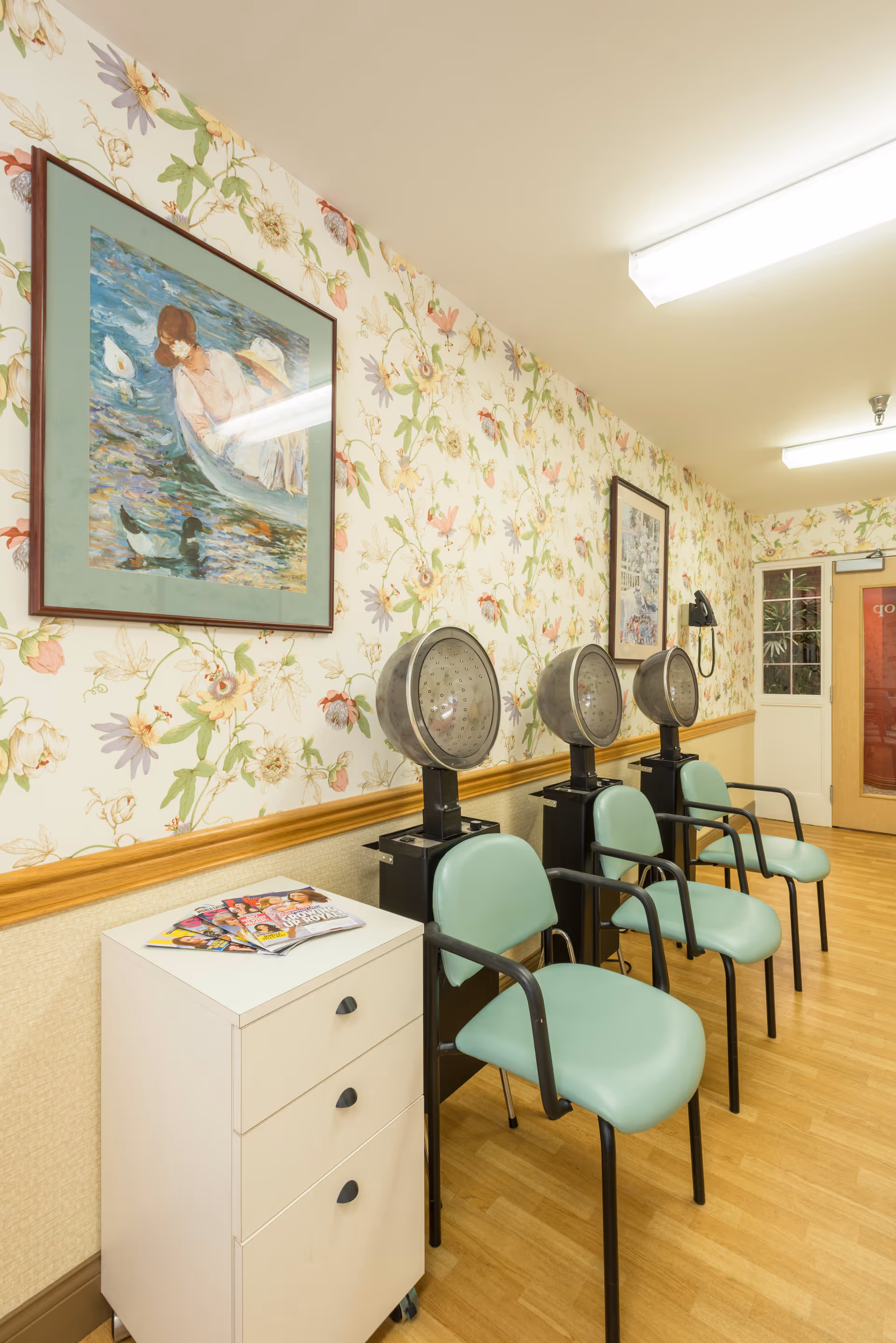 Row of vintage hair dryers with mint-green chairs against floral wallpaper in a salon area.