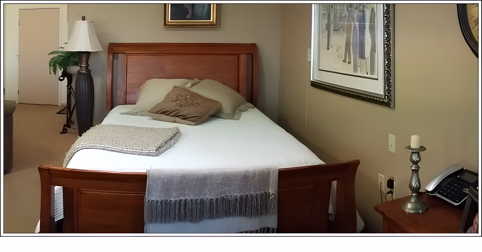 A cozy bedroom with a wooden bed frame, white bedding, and several pillows. A gray throw blanket is draped over the foot of the bed. To the right, there is a wooden nightstand with a silver candle holder and a telephone. The walls are beige with framed artwork hanging above the bed and on the side wall. A tall lamp and a green plant are visible in the background near a closed door.