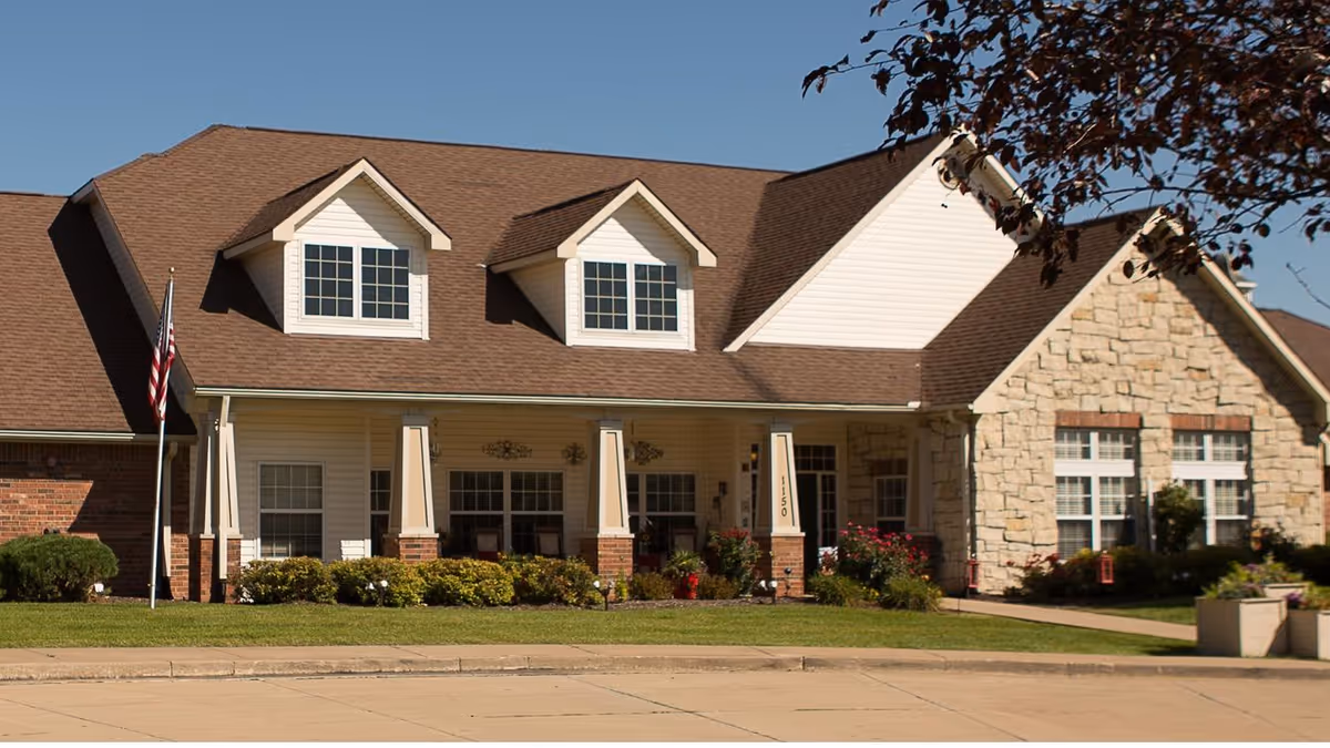 Front exterior view of a single-story senior living facility building with a brown shingled roof, white siding, stone accents, and a covered porch with columns. There is an American flag on a flagpole in the front yard, along with well-maintained bushes and flowers under a clear blue sky.