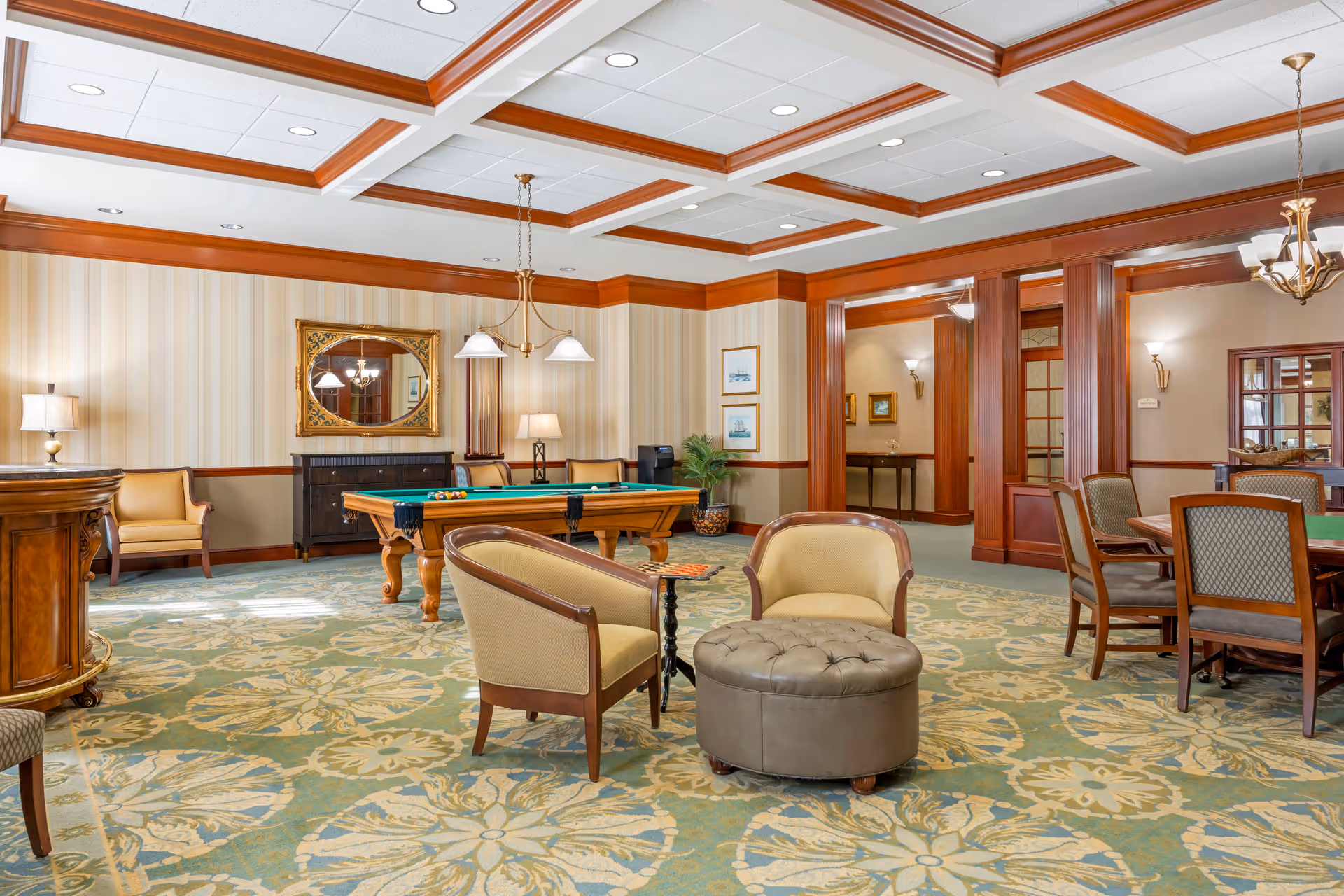 A spacious and elegant common area with a pool table, several chairs, a round ottoman, and a card table with chairs. The room features a patterned carpet, wood trim, and soft lighting from ceiling fixtures and lamps. There is a large mirror on one wall and framed artwork on another.