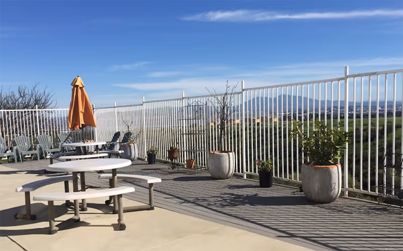 Sunlit outdoor patio with a white railing, potted plants, tables, chairs, and an umbrella.