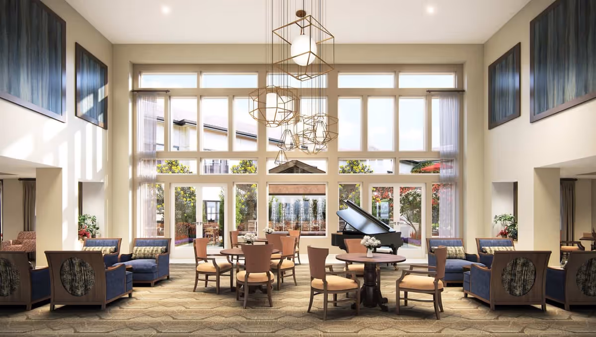 A spacious and well-lit living room area with large floor-to-ceiling windows allowing natural light to fill the space. The room features multiple round tables with chairs, several blue upholstered armchairs with patterned cushions, a black grand piano, and modern geometric pendant lights hanging from the ceiling. Outside the windows, a garden with trees and a water fountain is visible.