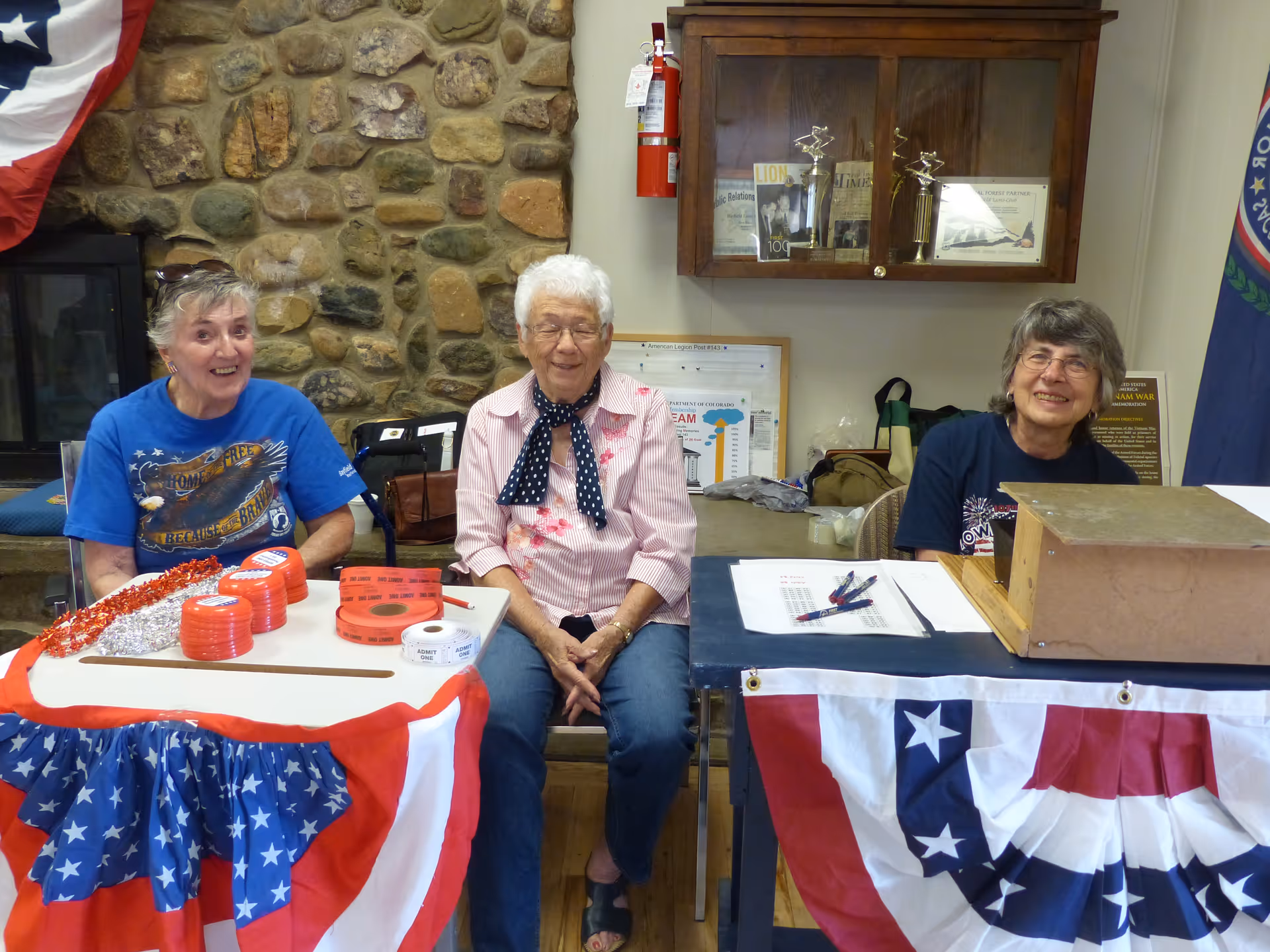Three elderly women sitting behind tables decorated with red, white, and blue patriotic bunting. The woman on the left is wearing a blue t-shirt and smiling, the woman in the middle is wearing a pink striped shirt with a navy polka dot scarf, and the woman on the right is wearing a dark blue t-shirt and glasses. Behind them is a stone wall, a wooden cabinet with trophies, and a fire extinguisher mounted on the wall.