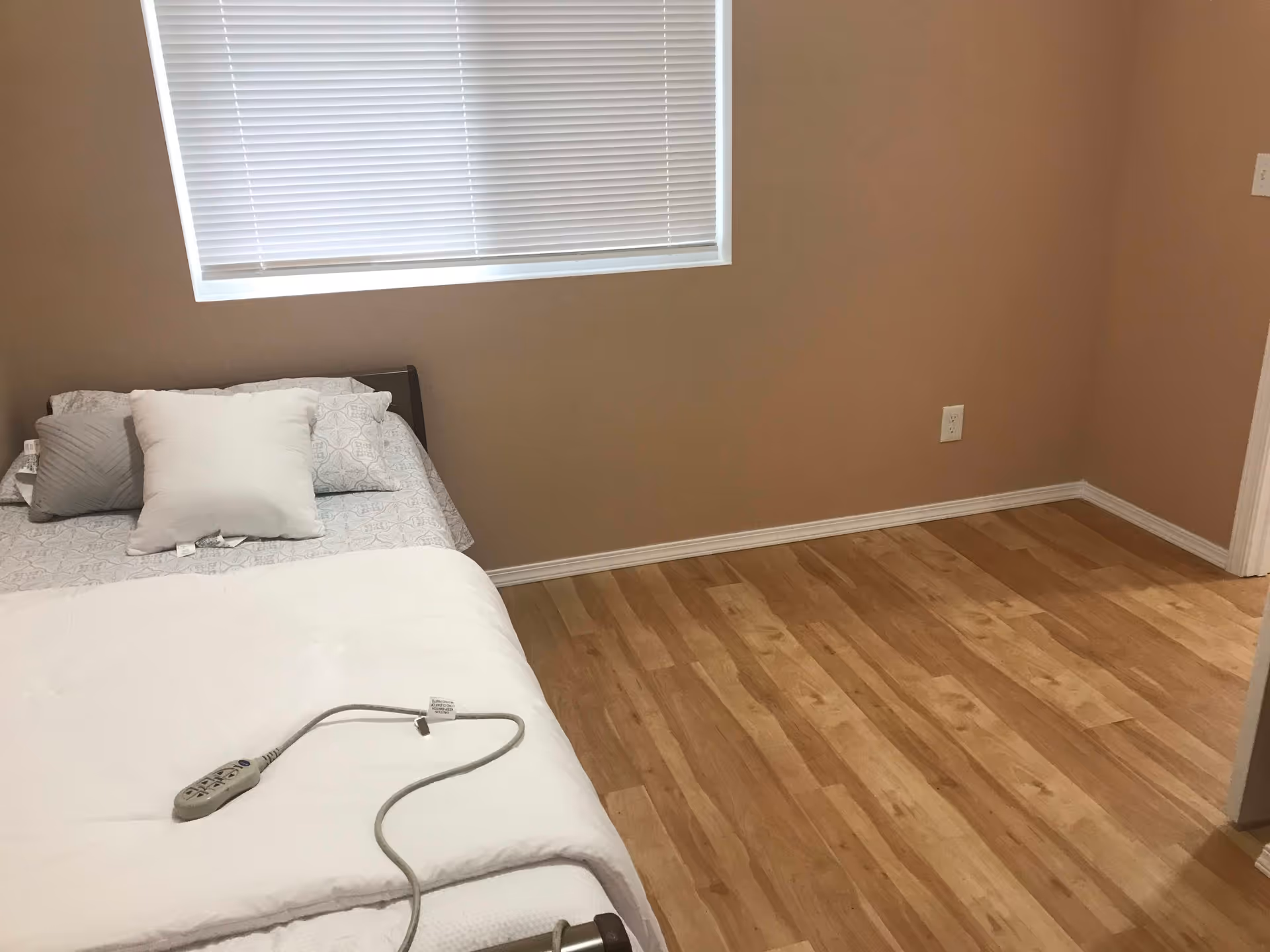 A simple bedroom with a bed against the wall under a window with closed blinds. The bed has a white comforter, patterned sheets, and several pillows. There is a remote control device on the bed, likely for adjusting the bed position. The room has light wood flooring and beige walls with electrical outlets visible.