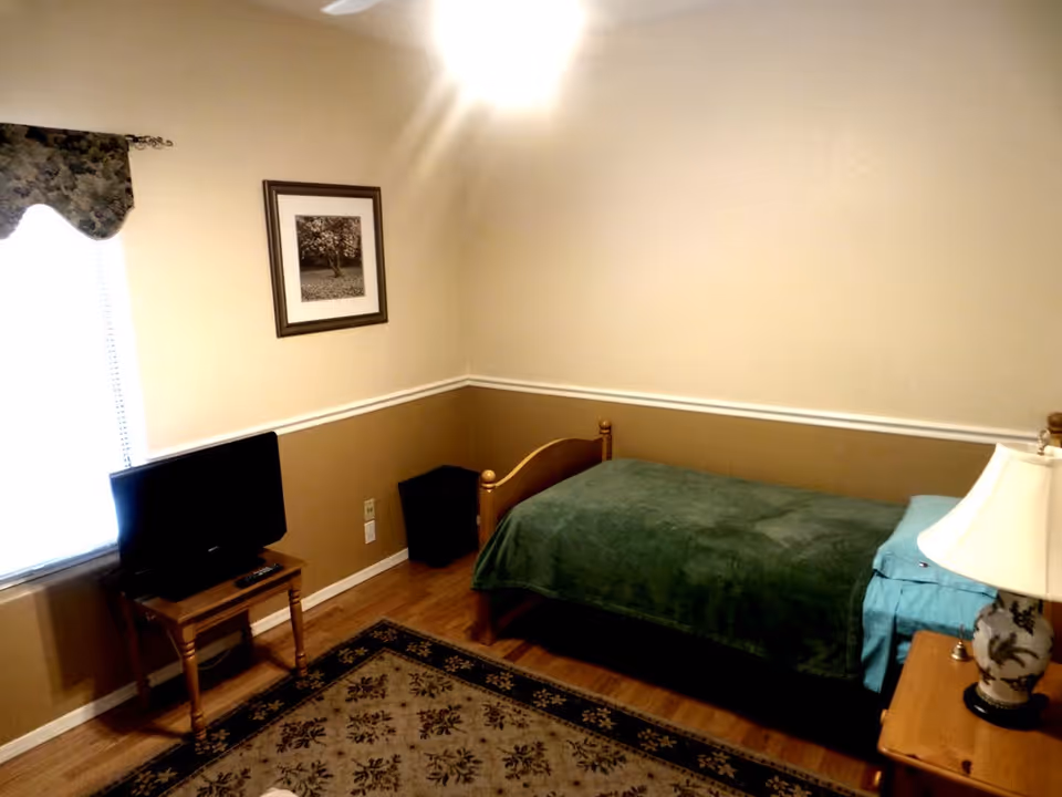 A small bedroom with a single wooden bed covered with a green blanket and blue pillow. There is a wooden nightstand with a decorative lamp on it next to the bed. A small wooden table holds a flat-screen TV near a window with a floral valance. A framed picture hangs on the beige and brown two-tone wall, and a patterned rug covers part of the wooden floor.