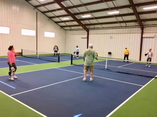 A group of six seniors playing pickleball indoors on a blue and green court with white lines. The facility has high ceilings with exposed beams and white walls with small windows near the ceiling.