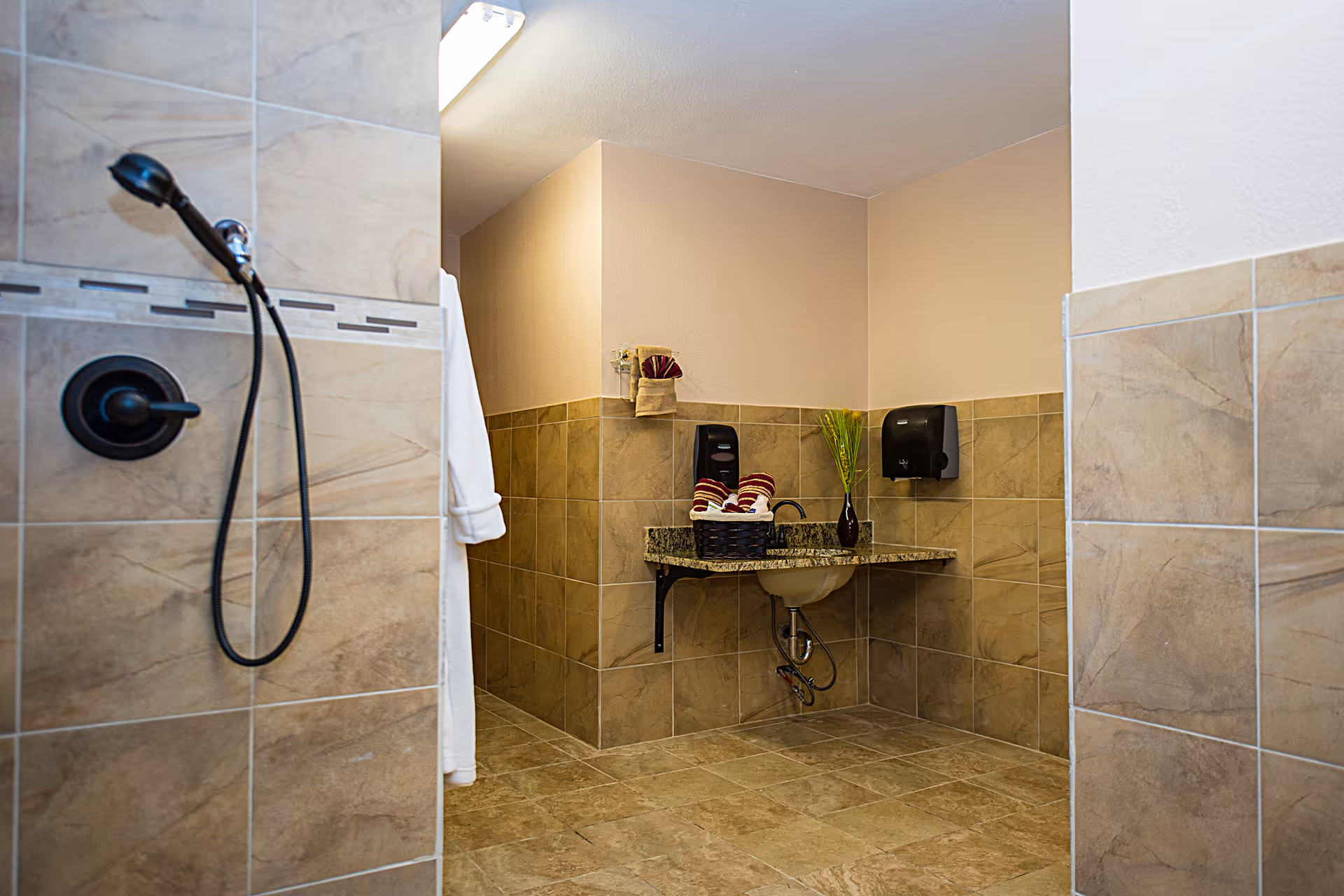 A clean and spacious bathroom area with beige tiled walls and floor. On the left, there is a black handheld showerhead attached to the wall. In the center, a granite countertop with a sink is mounted on the wall, holding a basket with rolled towels and a small vase with green decorative grass. A white robe hangs on the wall near the shower area, and there is a black soap or sanitizer dispenser mounted on the wall above the sink.