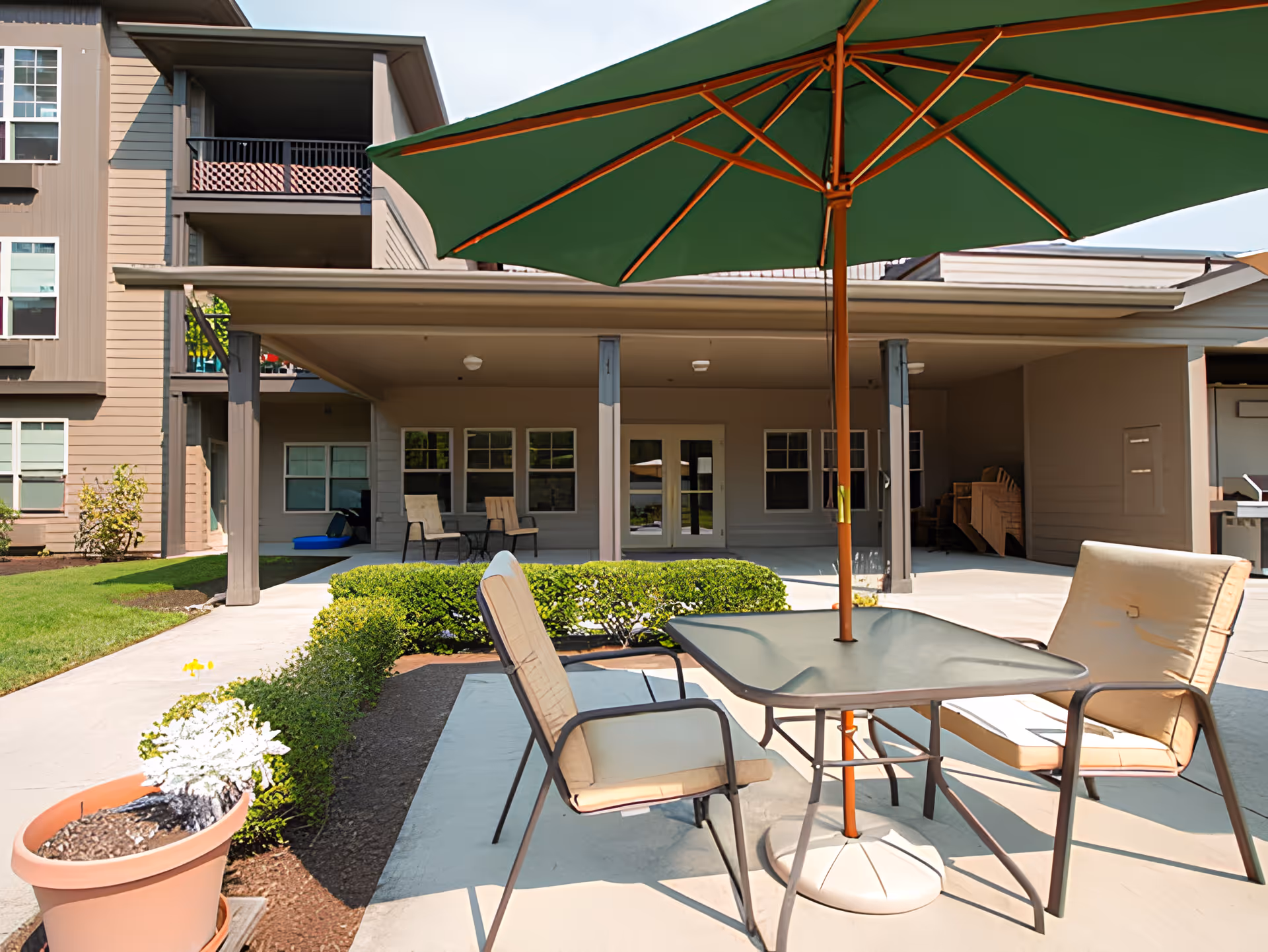 Outdoor patio area at Port Townsend Senior Living by Cogir featuring a table with a green umbrella and cushioned chairs, surrounded by neatly trimmed bushes and a potted plant, with the building's covered porch and windows in the background.