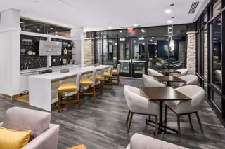 Spacious modern communal dining and lounge area featuring a long white island with stools, several small square tables with upholstered chairs, and glass entry doors.