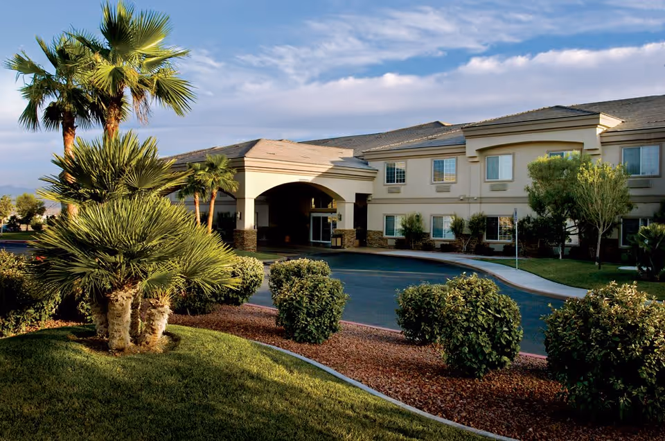 Exterior view of a senior living facility with a beige two-story building, an arched entrance, and a driveway. The foreground features green grass, palm trees, and neatly trimmed bushes under a partly cloudy sky.