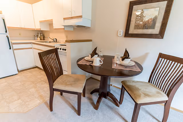 A small dining area with a round wooden table set for two, featuring plates, cups, and folded napkins. Two wooden chairs with cushioned seats are positioned around the table. In the background, there is a compact kitchen with white cabinets, a stove, and a refrigerator. A framed picture hangs on the wall above the table.