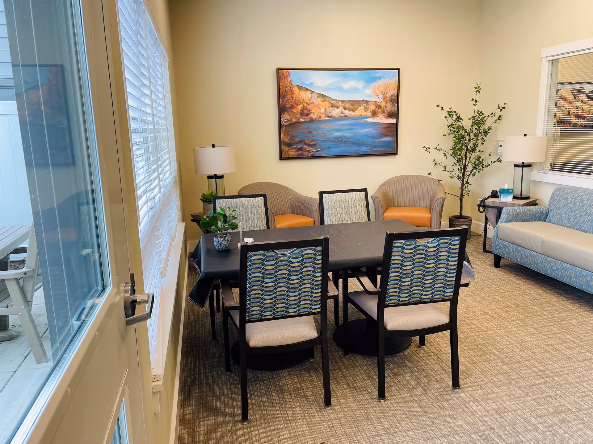 A cozy sitting area in a senior living community featuring a rectangular table with six chairs around it. The chairs have patterned backs and cushioned seats. Behind the table are two armchairs with orange cushions and a blue patterned sofa. The room is decorated with two table lamps, a potted plant, and a large framed painting of a river landscape on the wall. A window with blinds and a glass door leading outside are visible on the left side.