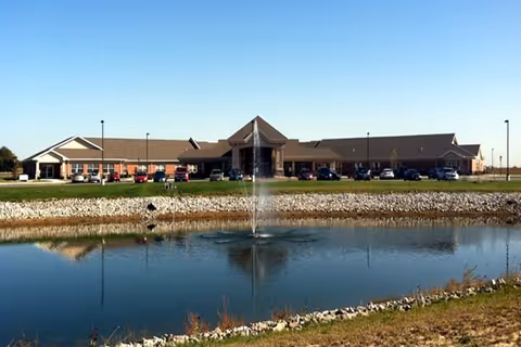 Front exterior of a single-story senior living building with a pond and fountain in the foreground.