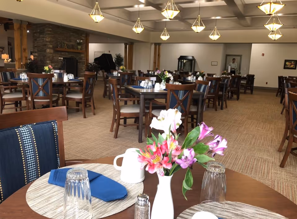 Spacious dining room with multiple set tables, floral centerpieces, pendant lights, and a stone fireplace in the background.
