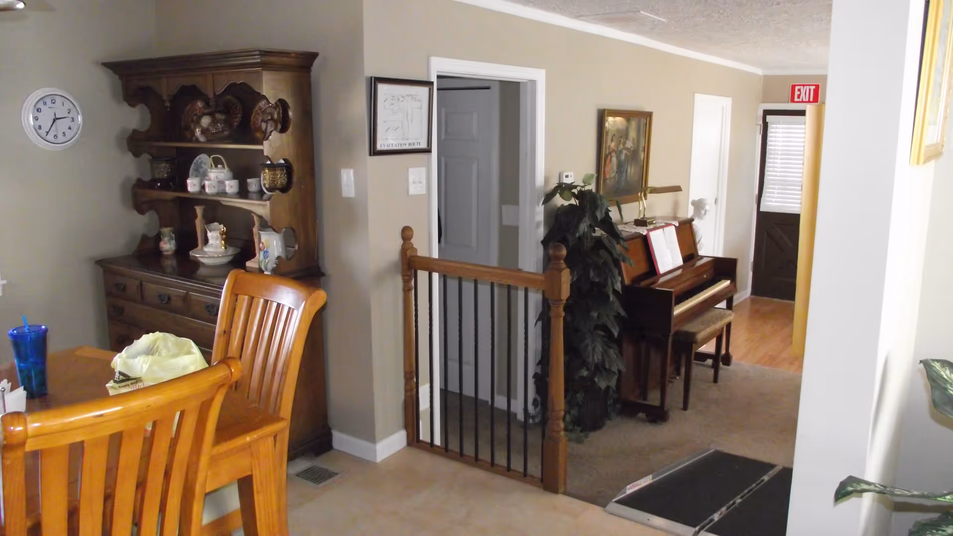 Interior view of a senior living facility showing a dining area with wooden chairs and a table, a wooden hutch with decorative items, a piano with a bench, a potted plant, and a hallway with an exit door. The walls are beige and the floor is a mix of tile and carpet.