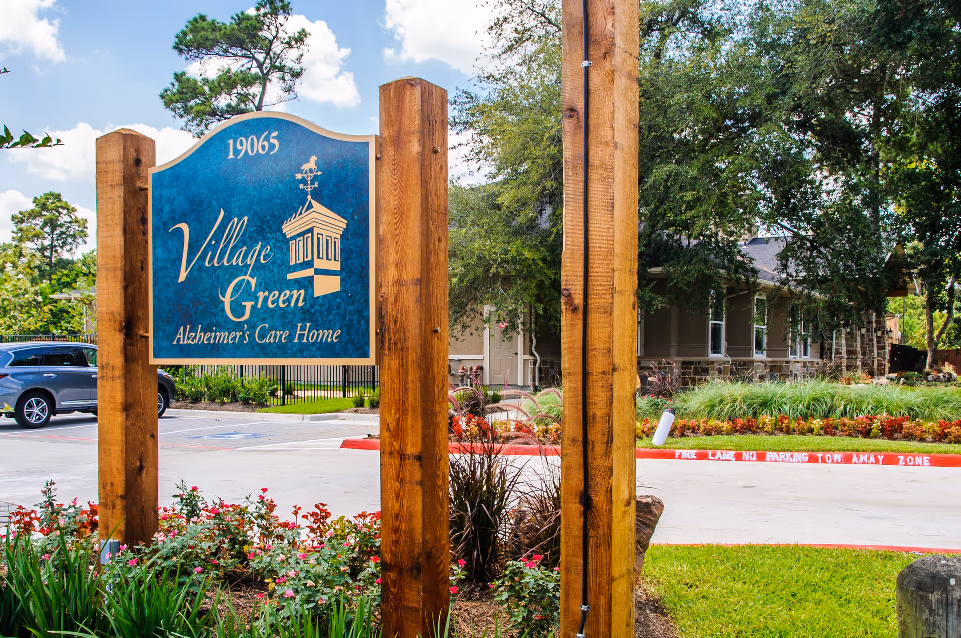 Wooden sign reading 'Village Green Alzheimer's Care Home' by a landscaped driveway in front of the facility building.