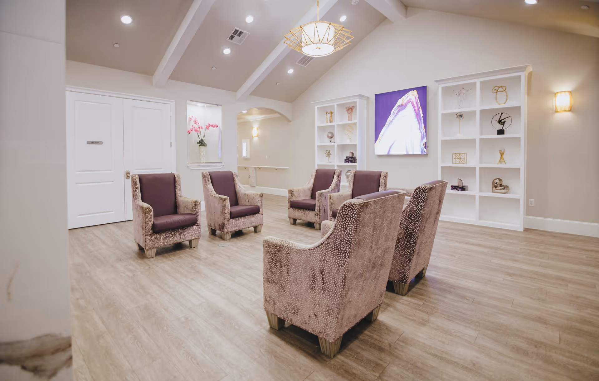 A bright and spacious common area with six upholstered armchairs arranged in a circle on a light wood floor. The room has high vaulted ceilings with recessed lighting and a modern chandelier. There are built-in white shelves on the far wall displaying decorative items, and a large abstract artwork with purple and white tones hangs between the shelves. A small window with pink flowers in a vase is visible on the left side of the room.