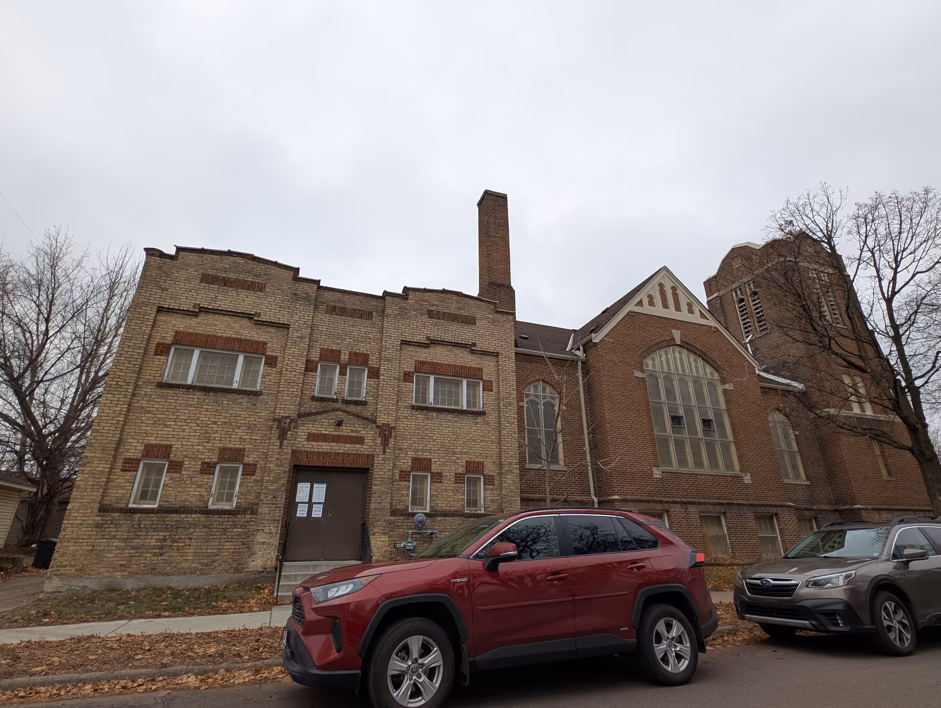 Exterior view of a large brick building with a tall chimney and arched windows, parked cars in front, and leafless trees on a cloudy day.