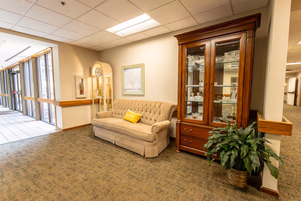 A cozy seating area in a senior living facility featuring a beige tufted sofa with a yellow pillow that says 'Together', a tall wooden grandfather clock, a wooden glass-front cabinet displaying crystal items, a framed picture of a tree on the wall, and a potted plant on the floor. The area is carpeted and adjacent to a hallway with large windows letting in natural light.