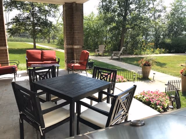 Outdoor patio area with a black square table surrounded by six black chairs with white cushions. There are two red cushioned chairs and a red cushioned loveseat near a brick column. The patio overlooks a grassy area with trees, a walking path, two beige Adirondack chairs, and flower beds with pink flowers.