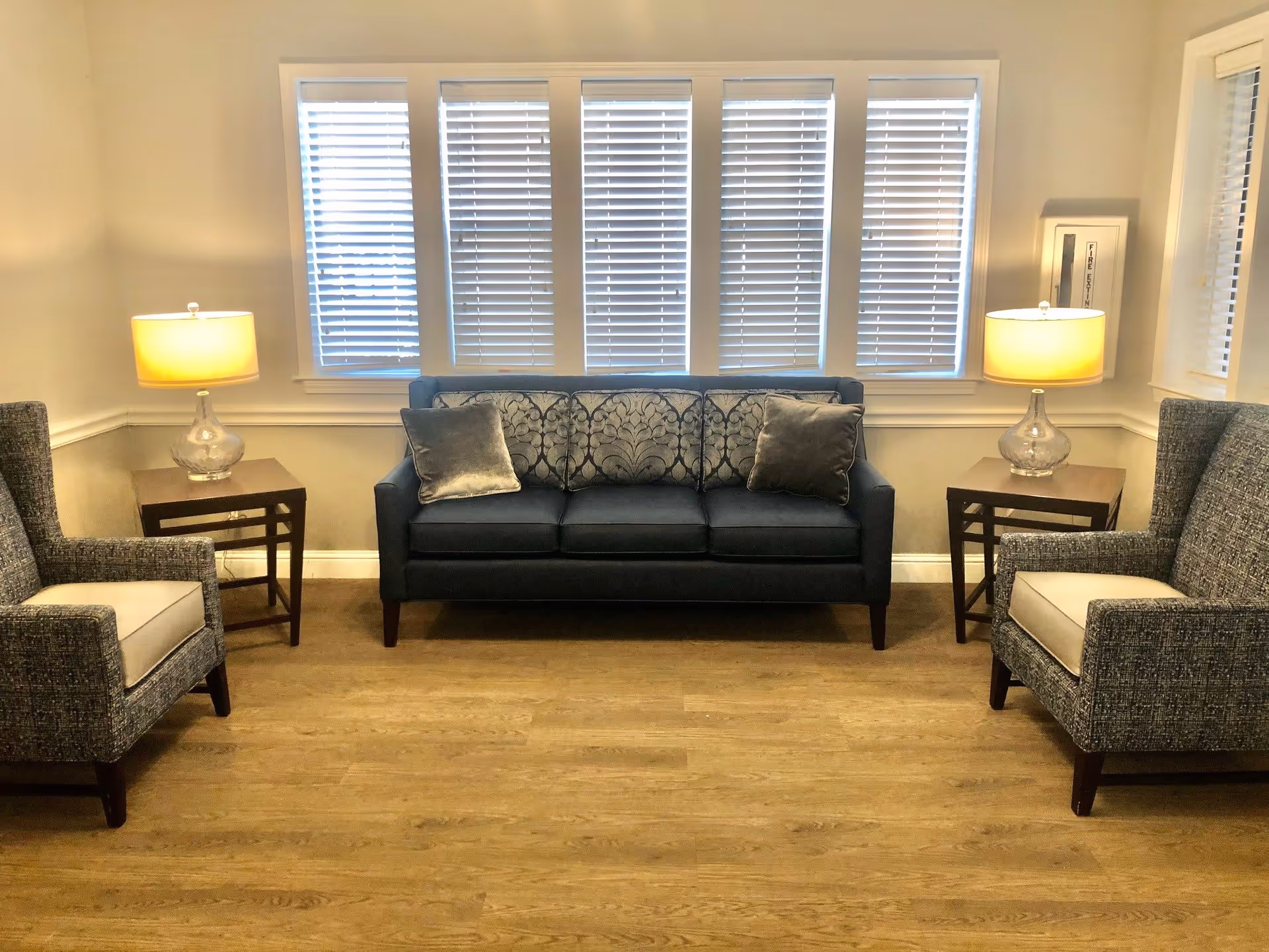 A cozy living room area with a dark blue sofa adorned with patterned and plain cushions, flanked by two matching armchairs with light cushions. Each side of the sofa has a wooden side table with a glass lamp on top. Behind the sofa are five windows with white blinds, and the room has light-colored walls and wooden flooring.