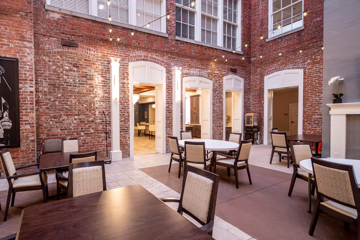 Indoor dining area with multiple tables and chairs arranged on a tiled floor, surrounded by exposed red brick walls with large windows above. String lights hang across the space, creating a warm and inviting atmosphere. Doorways lead to other rooms, and a fireplace with a white mantel and a potted orchid is visible on the right.