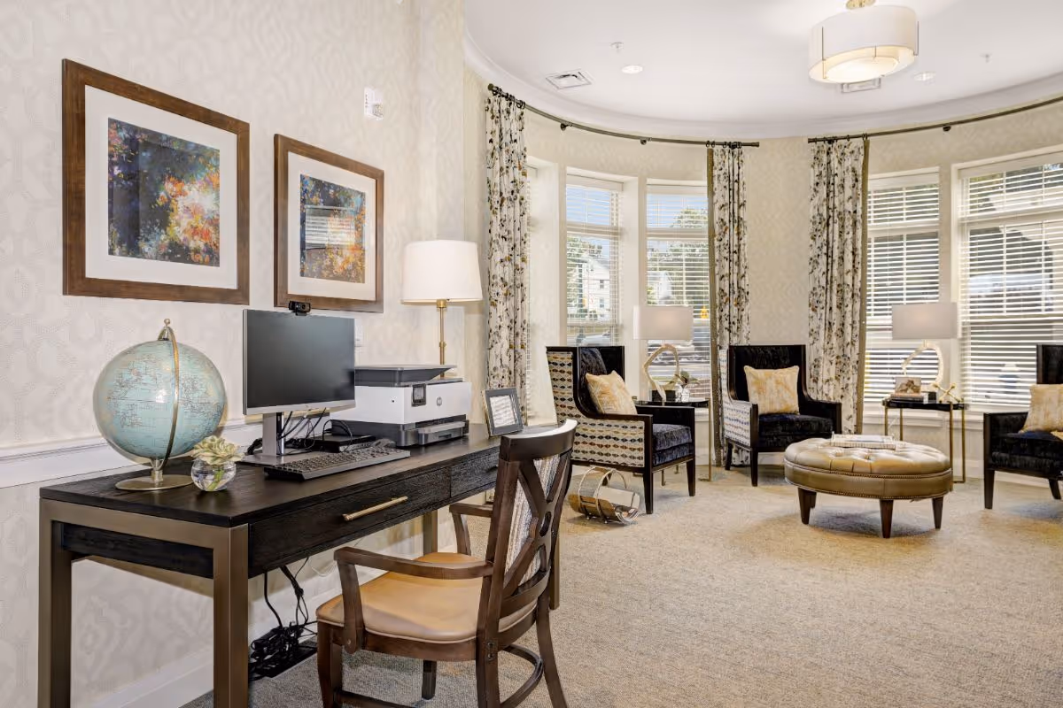 Bright senior living common room with a desk holding a computer and globe in the foreground and a seating area of armchairs and a round ottoman by bay windows.