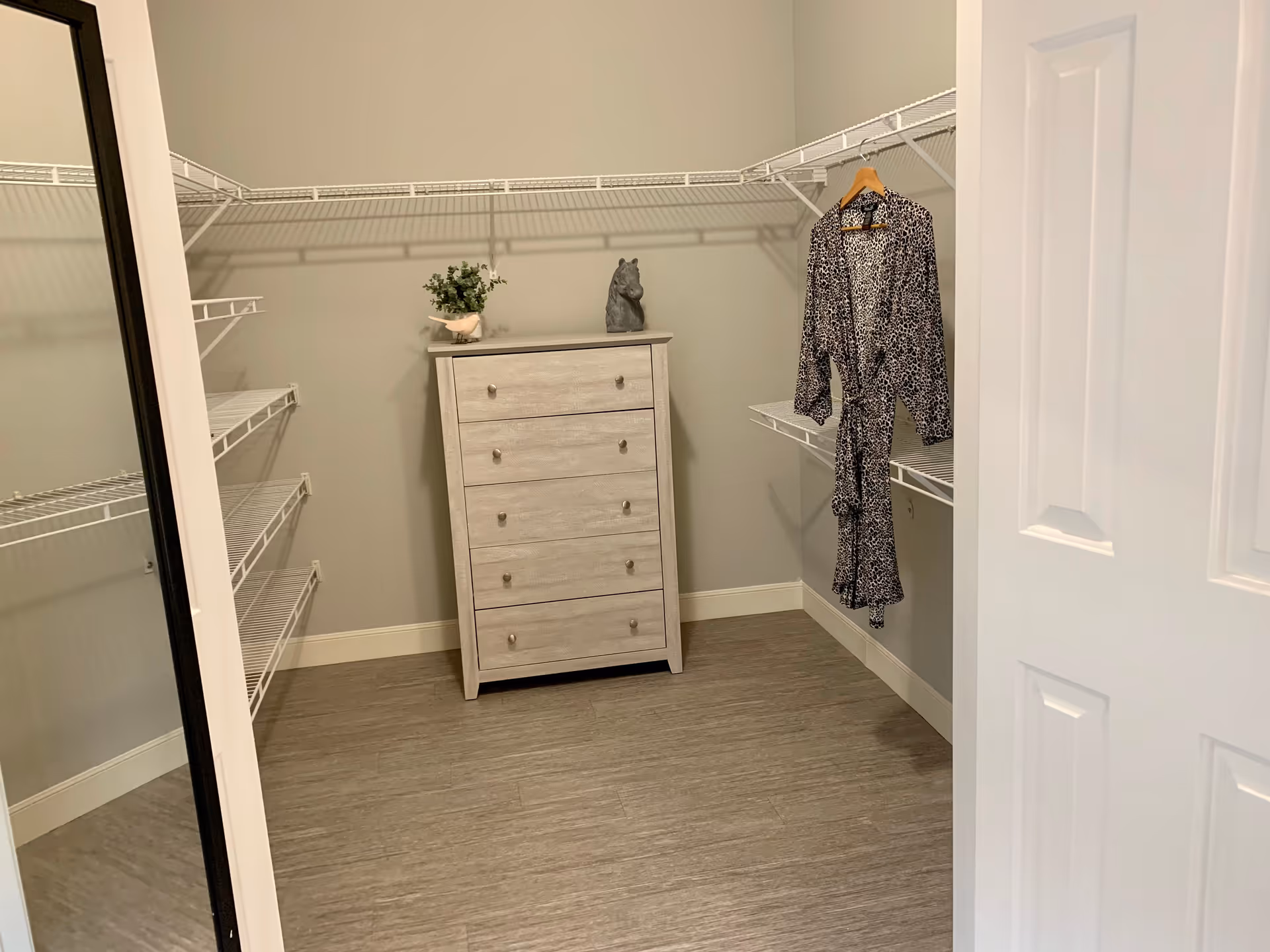 A walk-in closet with white wire shelving, a light wood dresser topped with small decor, and a patterned robe hanging on the right.