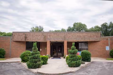 Brick facility entrance with glass doors, manicured shrubs, and a paved front walkway.