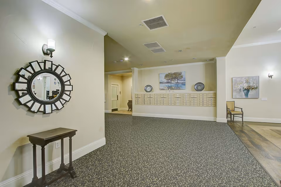Interior view of a senior living facility lobby area with a patterned carpet, a decorative round mirror above a small wooden table on the left wall, a row of mailboxes on the far wall beneath a painting of trees, and a single chair next to a floral painting on the right wall.