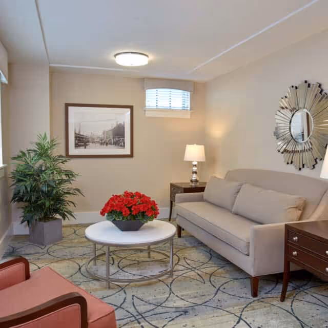 A cozy living room with a beige sofa, a round white coffee table with a red flower arrangement, a side table with a lamp, a decorative mirror on the wall, a framed black and white photo, a potted plant, and an armchair with wooden arms.