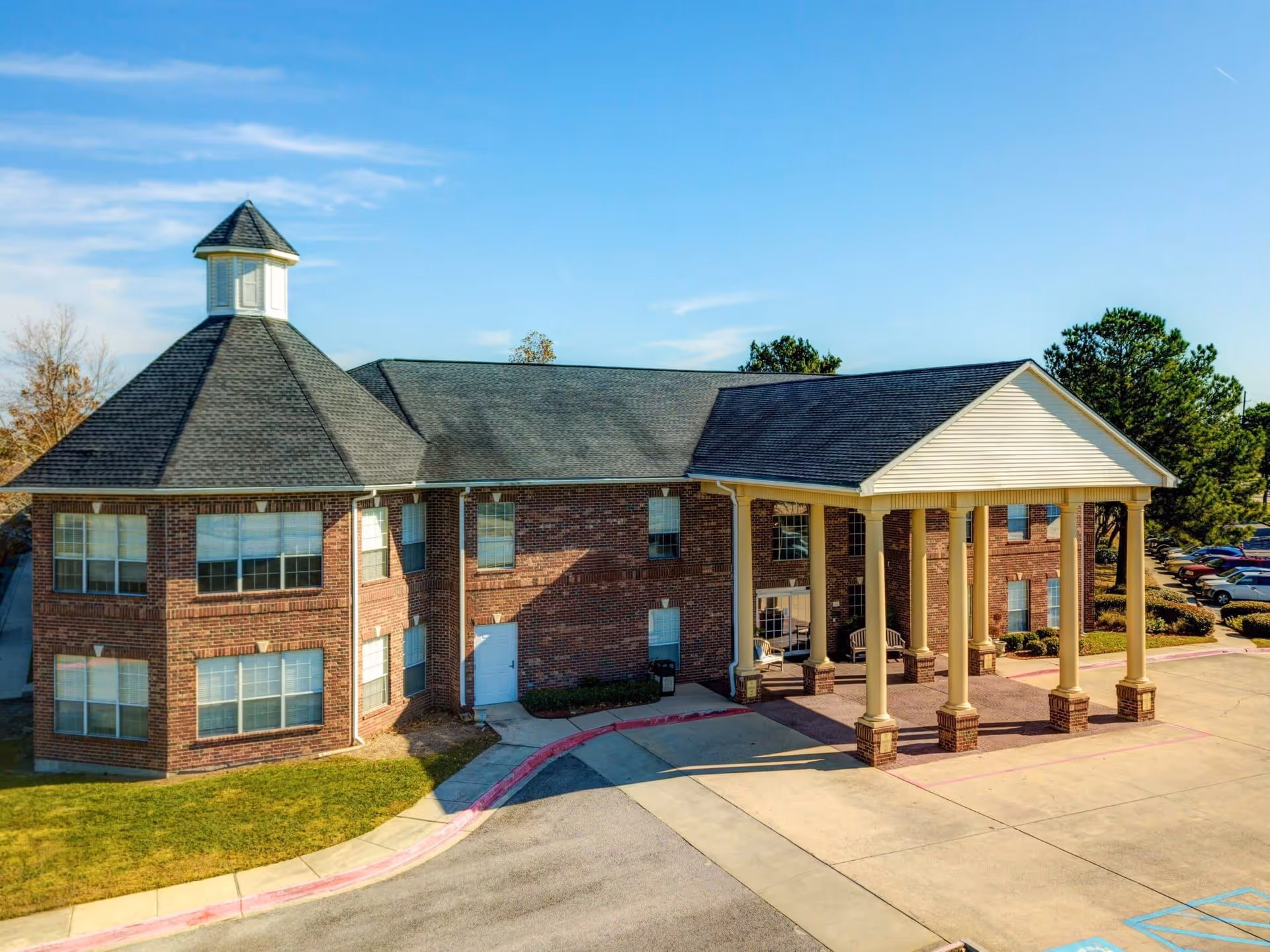 Exterior view of a two-story brick building with a large covered entrance supported by six columns, surrounded by a parking area and some trees under a clear blue sky.