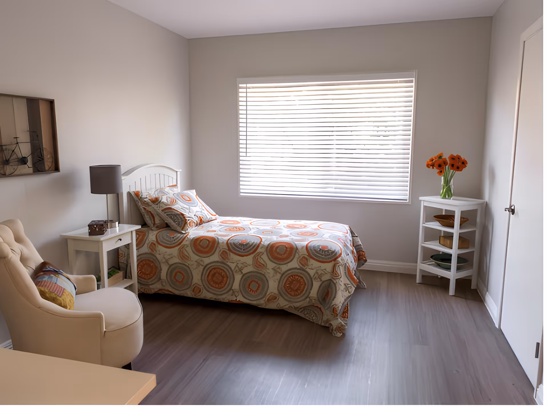 A bright single bedroom with a patterned bed, armchair, nightstand and lamp, a window with blinds, and a small shelving unit with flowers.