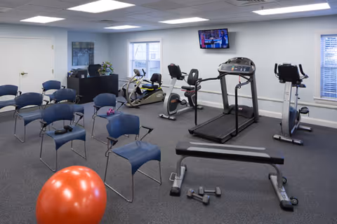 A fitness room in a senior living facility with exercise equipment including a treadmill, stationary bikes, a bench, and dumbbells. Several blue chairs are arranged in rows, and a large orange exercise ball is in the foreground. A television is mounted on the wall, and windows allow natural light into the room.