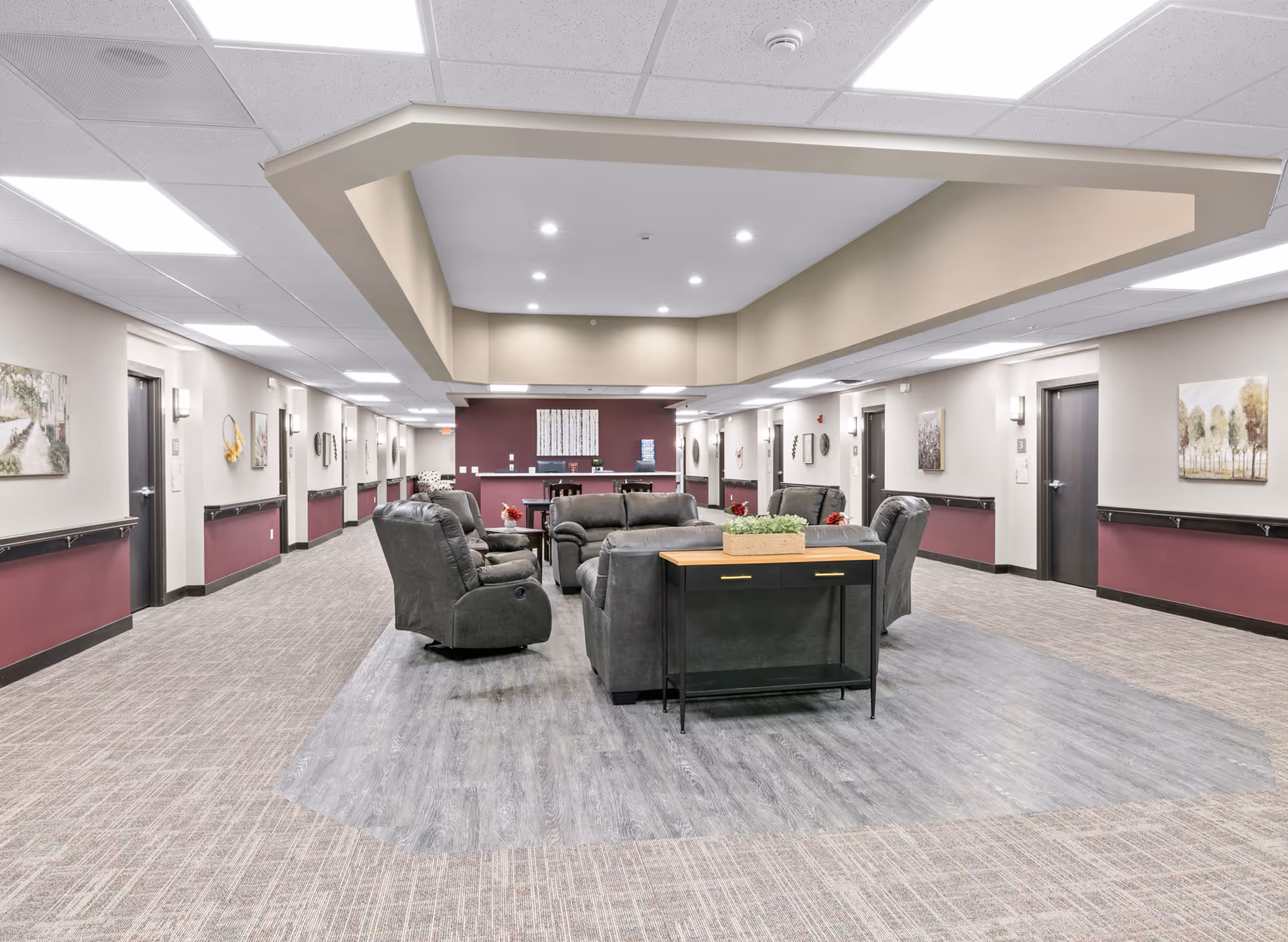 Open communal lounge in a senior living facility with recliner chairs and sofas arranged under a recessed ceiling in a wide hallway.