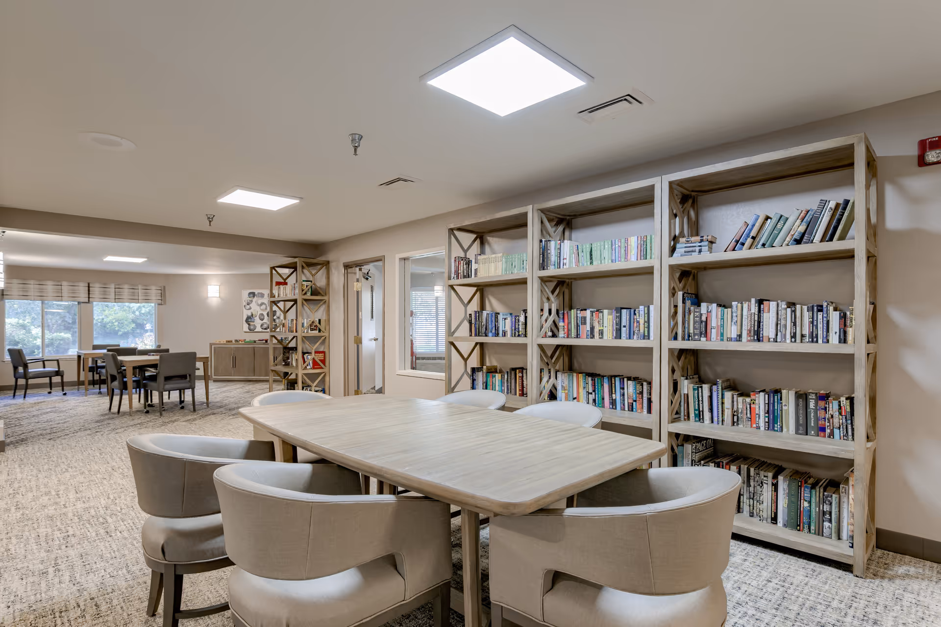 A cozy interior room with a wooden table surrounded by six cushioned chairs in the foreground. Behind the table, there are three large wooden bookshelves filled with books. In the background, there is another seating area with tables and chairs near large windows letting in natural light. The room has a neutral color palette and carpeted floors.