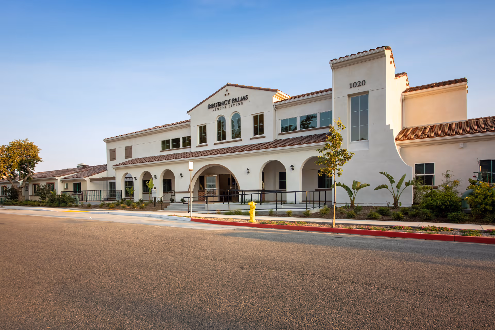 Exterior front view of Regency Palms Senior Living facility in Oxnard, featuring a two-story building with white stucco walls, arched entryways, and a tiled roof under a clear blue sky.