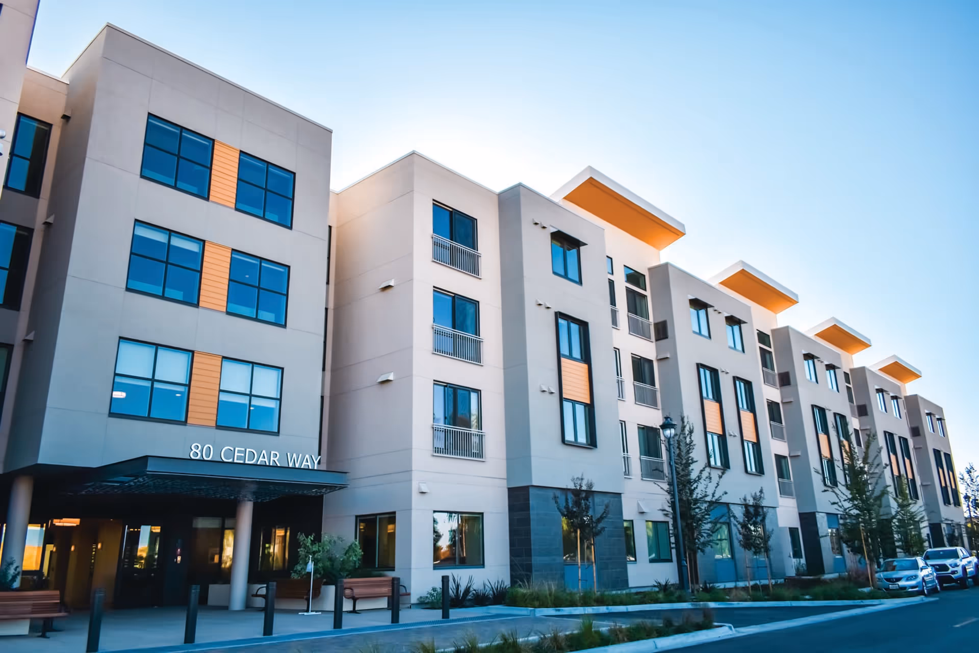 Exterior view of a modern multi-story building with large windows and orange accents, featuring the address 80 Cedar Way. There are benches and small trees near the entrance, and several cars parked along the street.