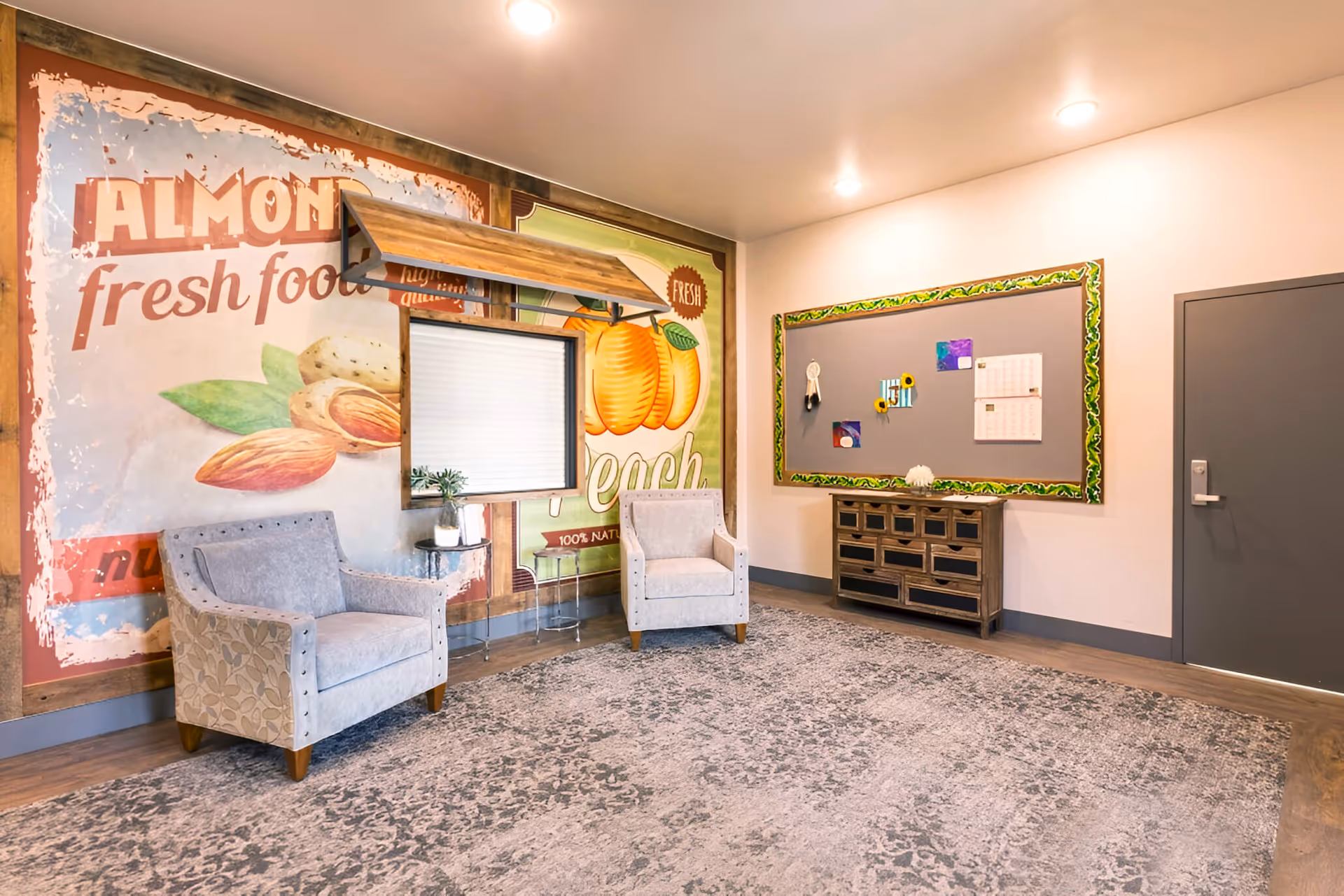 A cozy interior seating area with two upholstered armchairs placed on a patterned rug. Behind the chairs is a large wall mural featuring illustrations of almonds and peaches with text promoting fresh food. To the right, there is a bulletin board with a decorative green border, some pinned notes, and a wooden cabinet with multiple drawers beneath it. A closed gray door is visible on the far right wall.