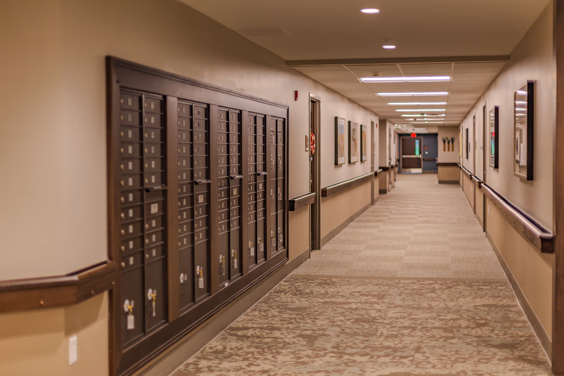 Long carpeted interior hallway in a senior living facility with wall-mounted mailboxes, handrails, and framed artwork.