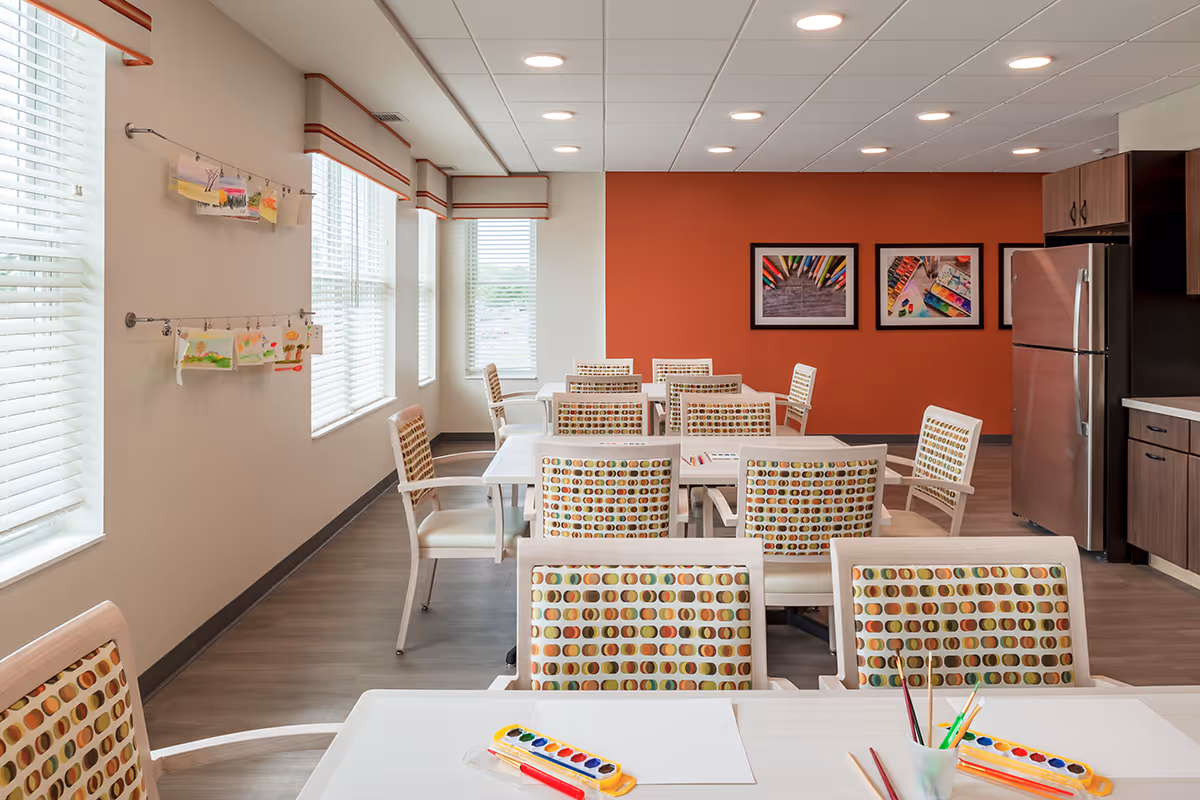A bright and clean activity room with multiple tables and chairs arranged neatly. The chairs have patterned upholstery with circular designs. On the tables are watercolor paint sets, brushes, and blank sheets of paper, suggesting an art activity setup. The room has large windows with blinds letting in natural light, an orange accent wall with framed colorful artwork, and a kitchenette area with a stainless steel refrigerator and wooden cabinets.