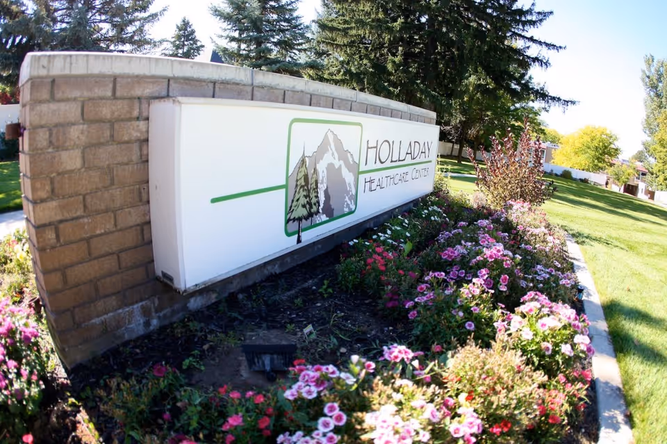 Outdoor view of a brick and white sign for Holladay Healthcare Center surrounded by a flower bed with pink and purple flowers, green grass, and trees in the background under a clear blue sky.